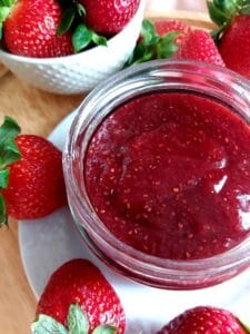 Jar filled with strawberry jam on a white plate garnished with fresh strawberries