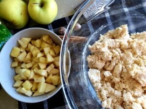 A mixing bowl with dough, a small bowl with sliced apples, an apple next to the bowl and a cinnamon stick.
