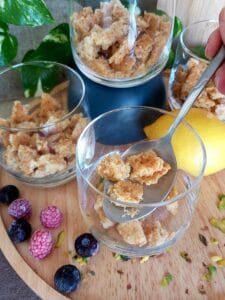 Crumbled sponge cake in glass dessert cups, ready for layering with fruit curds and pistachio cream. Fresh blueberries, raspberries, and chopped pistachios are scattered on a wooden board, with lemons in the background.